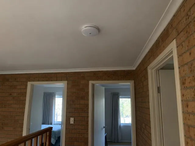 Ceiling-mounted smoke alarm above a brick-walled hallway with doorways and stair railing in Erina